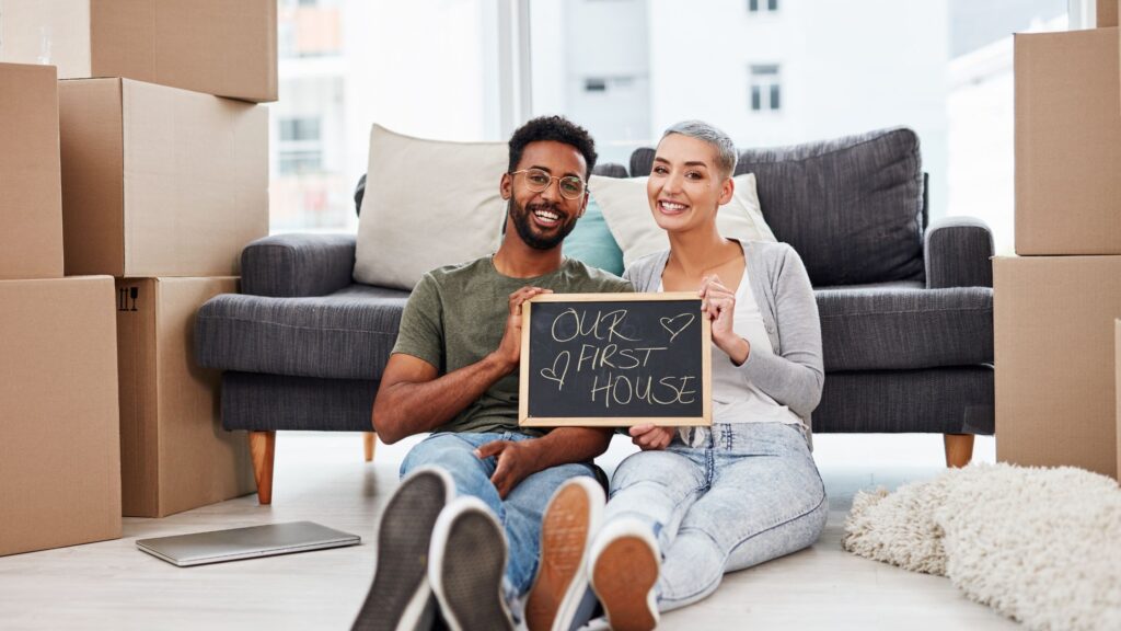 A couple sits in a room with a sign that says our first home